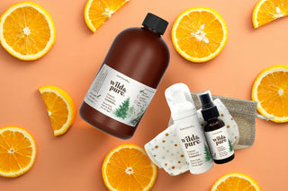 A flat-lay product arrangement on a peach background with orange slices around the edges. The set includes an EcoBalance Cleanser 18 fl oz refill and an On-the-Go Pack set of cleansers in mist and foam with organic cotton flannel dry wipes plus a burlap bag to carry the travel set.