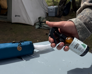 Person holding Wild & Pure Natural 4oz EcoBalance Cleanser at a camp site with Wild & Pure 100% cotton dry wipes in a dispenser on the table nearby.