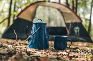 Blue enamel coffee pot and mug on the ground in front of a tent in a forest.