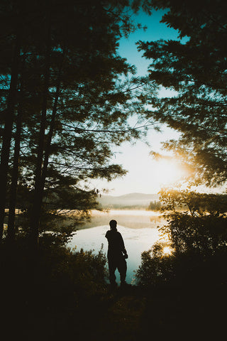 Person standing by a lake with trees and sunset in the background